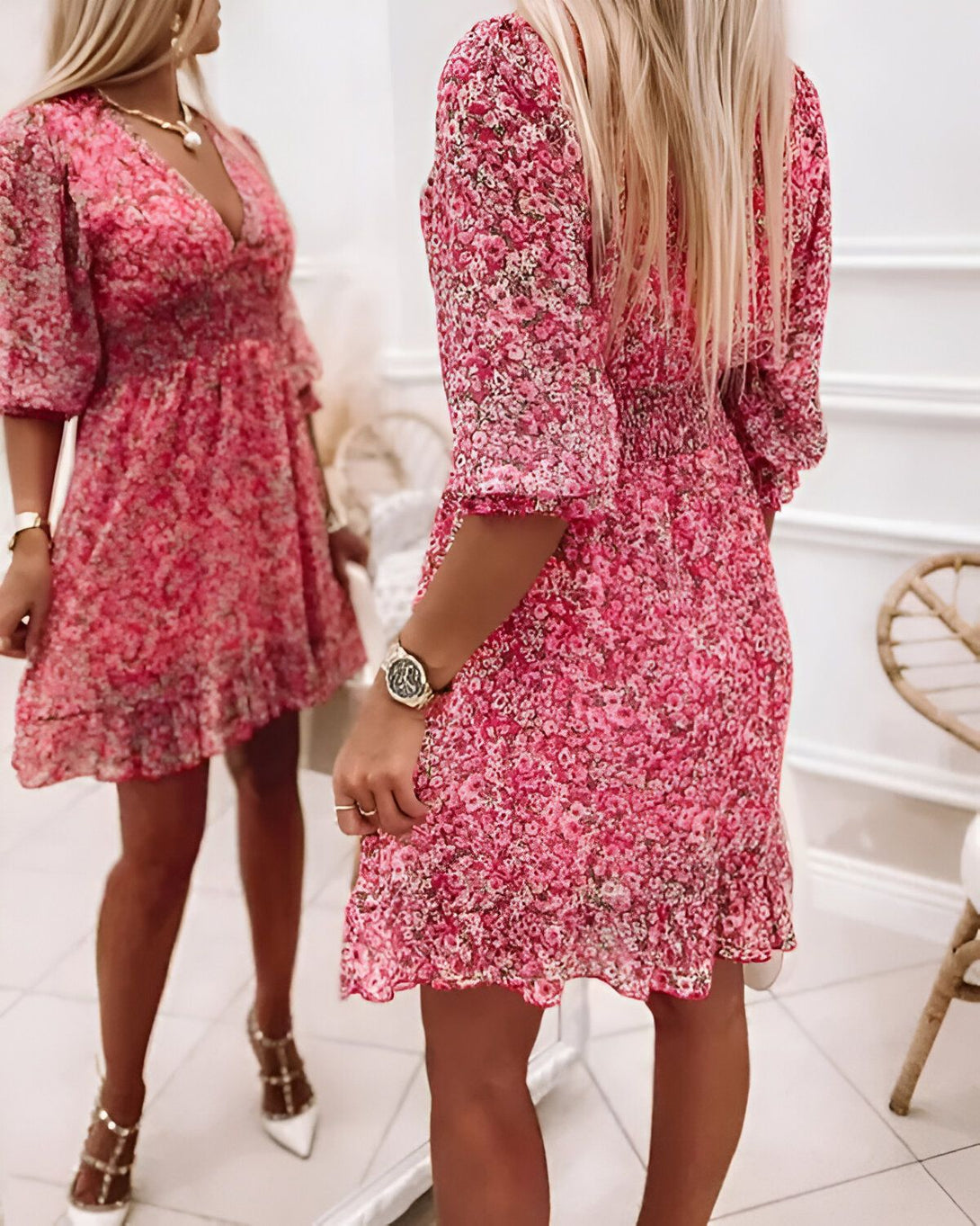 Une jeune femme porte une robe rose à fleurs. La robe est courte et évasée, avec des manches bouffantes. La femme est blonde et porte des talons hauts blancs. Elle est debout devant un miroir et sourit à son reflet.