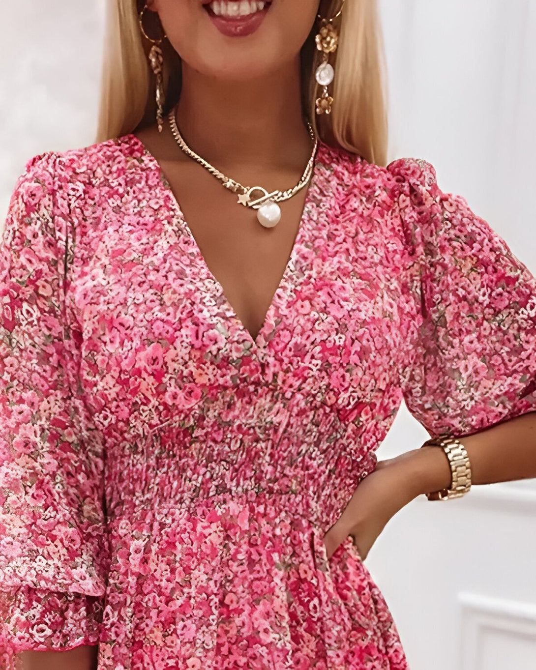 Une femme porte une robe rose à fleurs avec un décolleté en V et des manches bouffantes. Elle porte également un collier avec un pendentif en perle et des boucles d'oreilles en perles.
