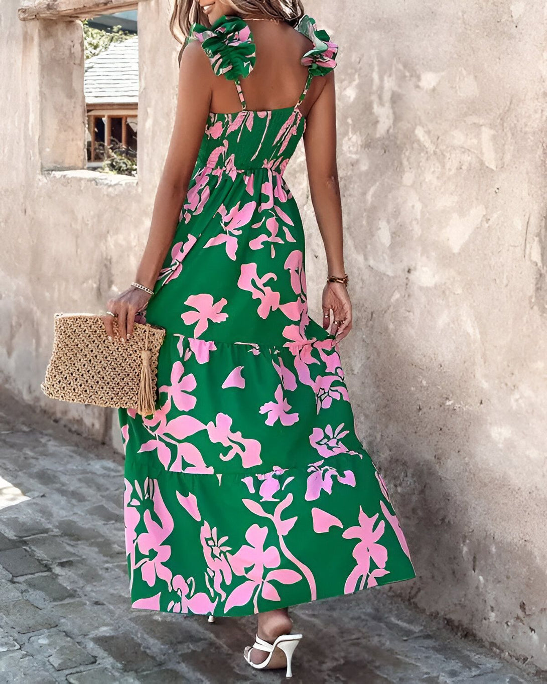 Une femme porte une robe longue verte à motifs floraux roses. La robe est froncée à la taille et possède des bretelles fines. La femme porte également un sac à main en paille marron et des sandales blanches à talons hauts.