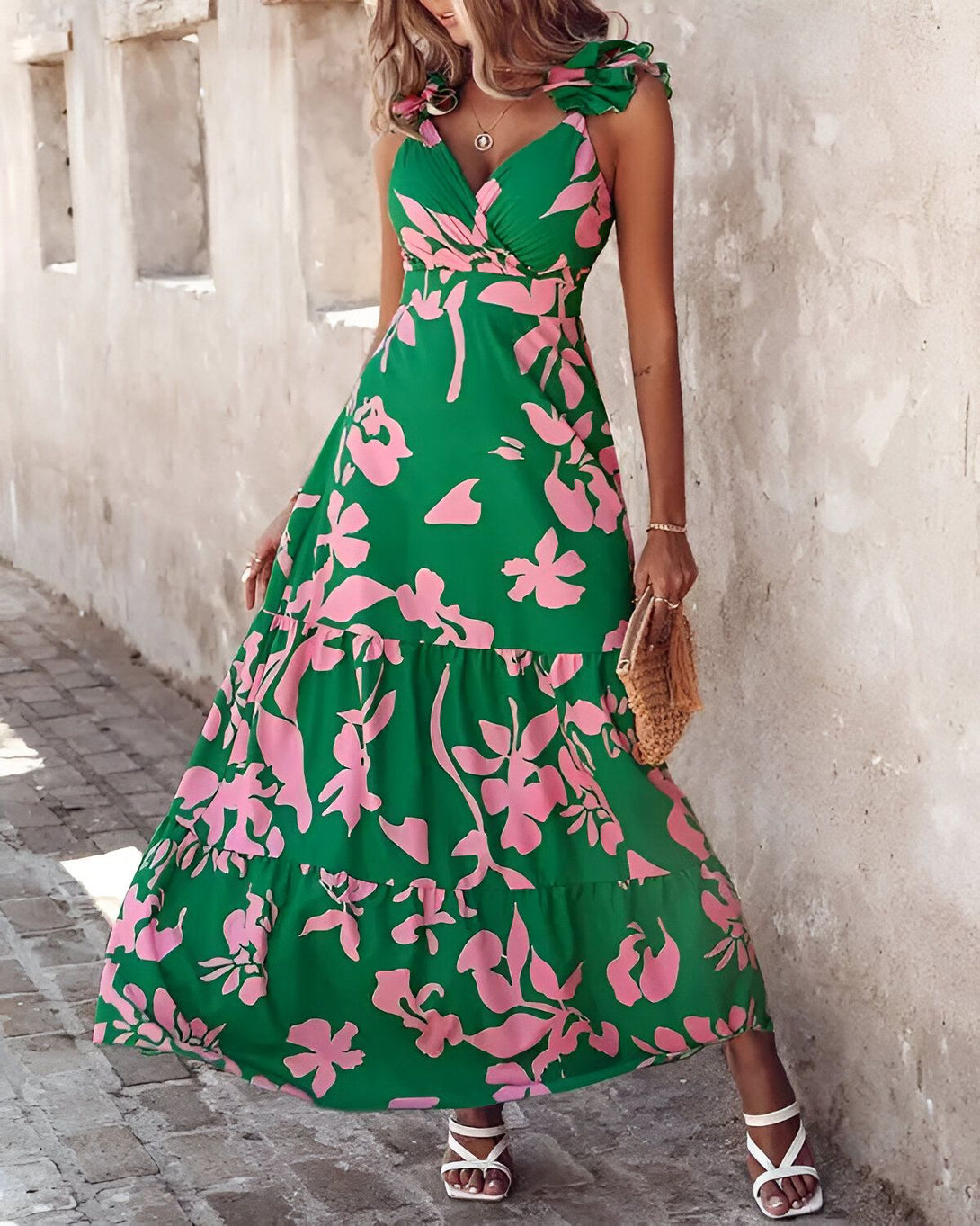 Une femme porte une robe longue verte à motifs floraux roses. La robe est décolletée et a des bretelles fines. La femme est debout dans une rue et porte des sandales blanches.