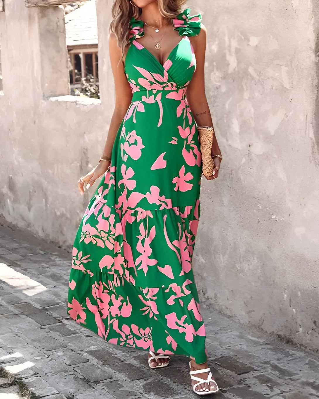 Une femme porte une robe longue verte à motifs floraux roses. La robe est décolletée et possède des bretelles fines. La femme est debout dans une rue et porte des sandales blanches.