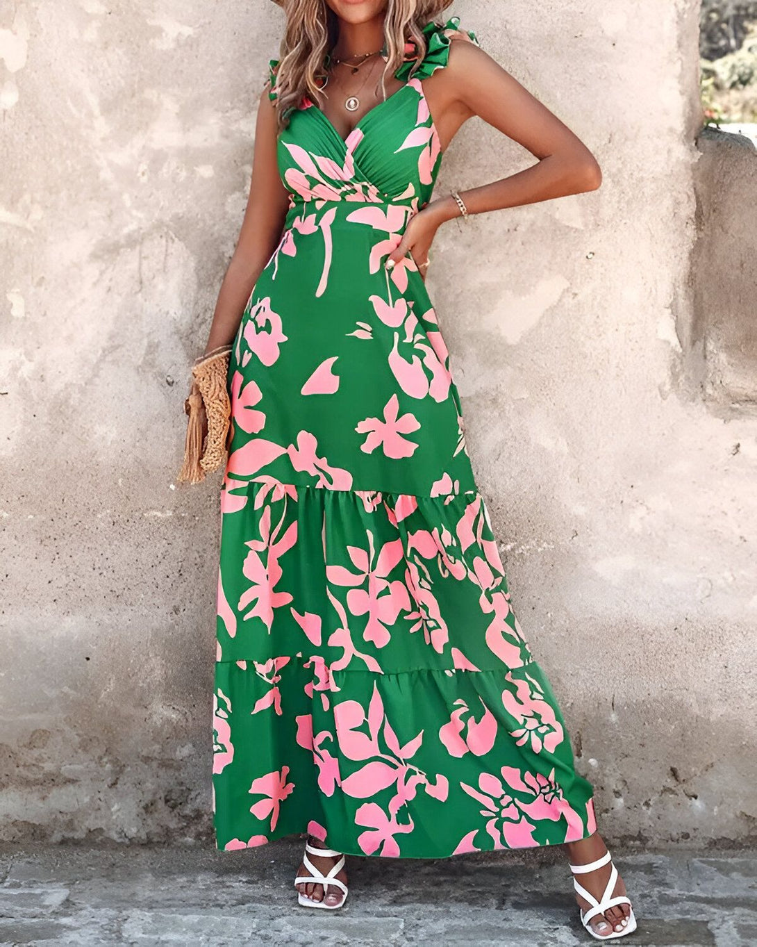 Une femme porte une robe longue verte à motifs floraux roses. La robe est décolletée et possède des bretelles fines. La femme est debout, adossée à un mur en pierre. Elle porte des sandales blanches et un sac à main en osier.