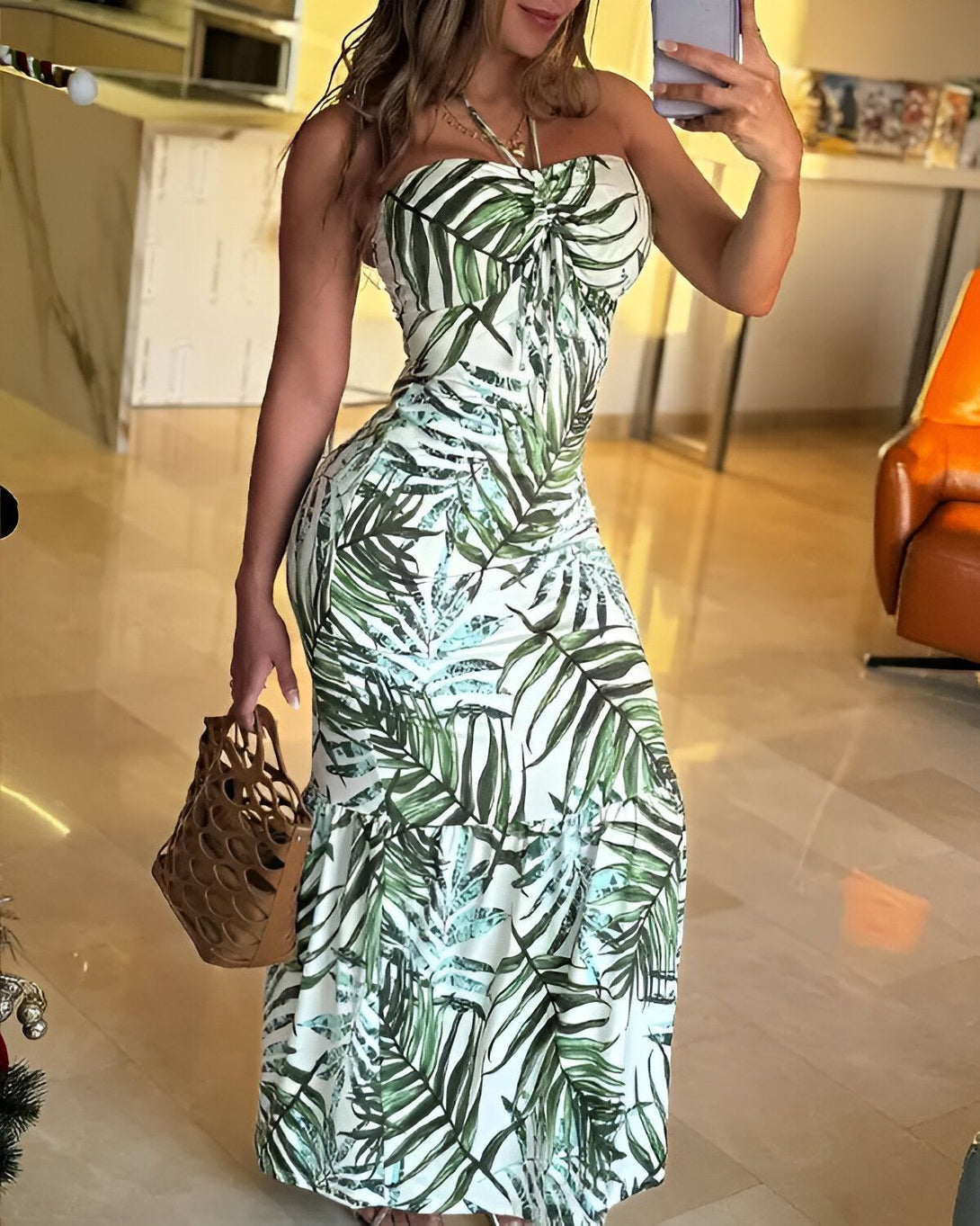 Une femme porte une robe longue blanche et verte à motifs de feuilles de palmier. Elle est debout dans un salon et tient un sac à main marron.