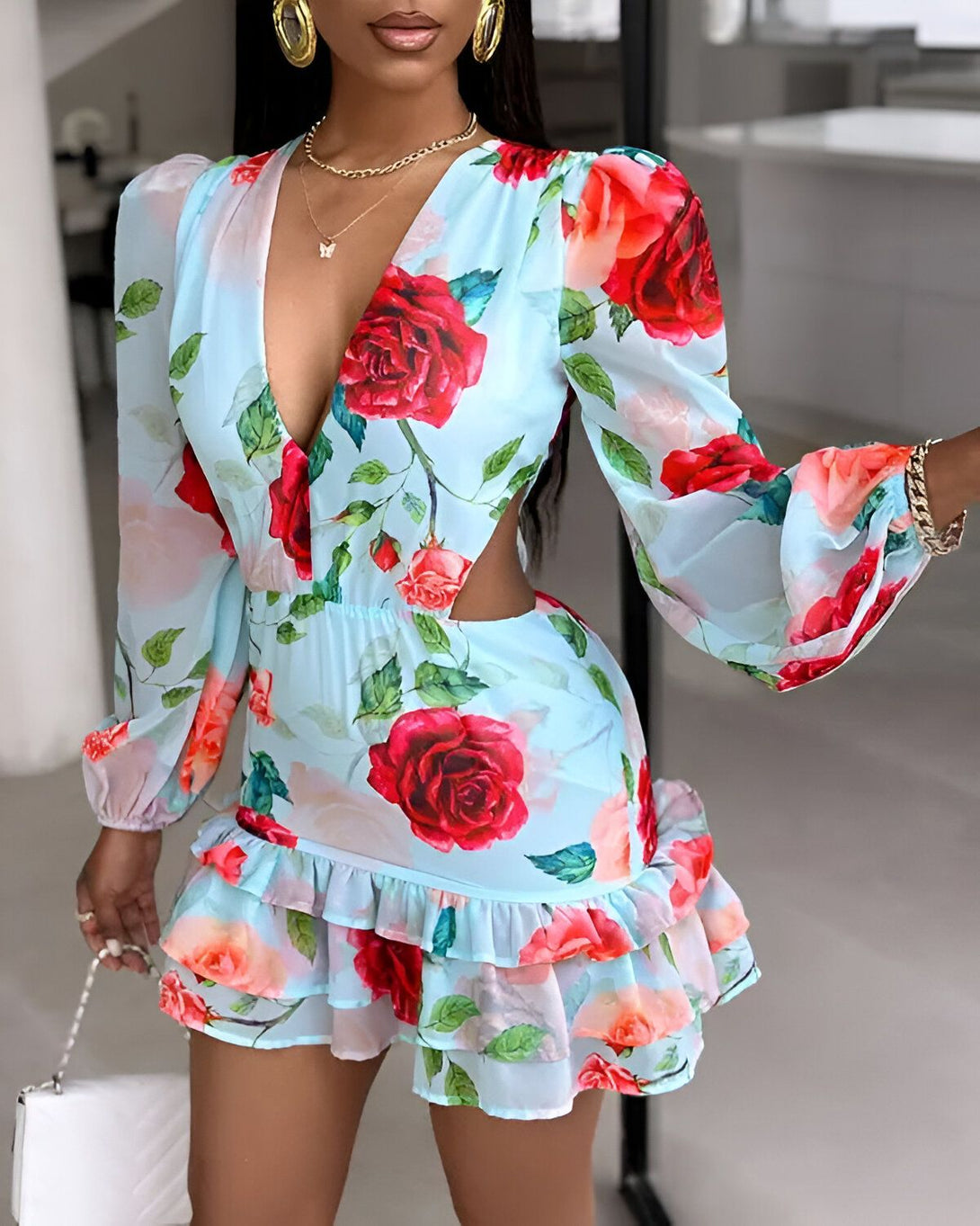Une femme porte une robe bleu clair à motifs de roses rouges. La robe est courte et possède des manches longues. Elle est cintrée à la taille et possède un décolleté en V. La femme porte également des escarpins rouges et un collier doré.