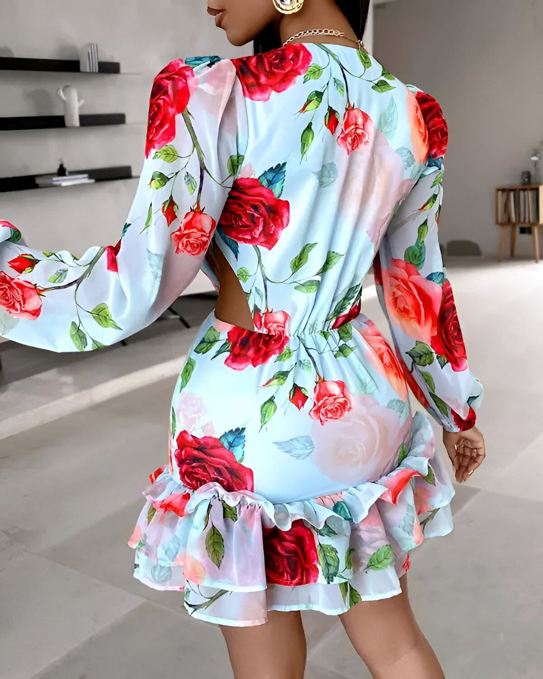 Une femme porte une robe bleu clair à motifs de roses rouges et roses. La robe est courte et possède des manches longues. Elle est cintrée à la taille et possède un décolleté plongeant dans le dos. La femme est vue de dos et porte des talons hauts couleur chair.