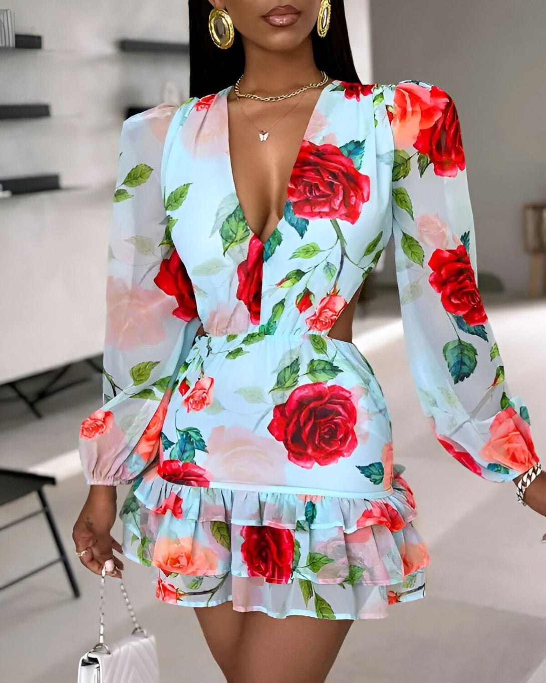 Une femme porte une robe bleu clair à fleurs rouges et roses. La robe est courte et a des manches longues. Elle est cintrée à la taille et a un décolleté en V. La femme est debout dans une pièce et porte des talons hauts blancs.