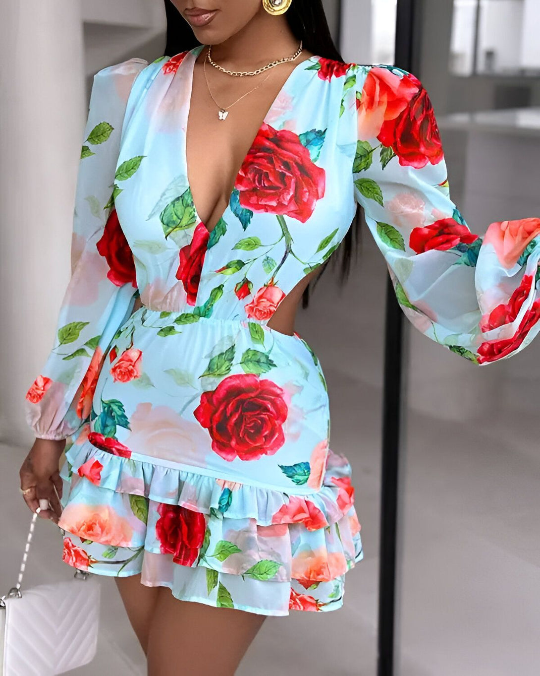 Une femme porte une robe bleu clair à motifs de roses rouges et de feuilles vertes. La robe est courte et possède des volants sur le bas. Les manches sont longues et bouffantes. La femme est debout, elle porte un collier et un sac à main blanc.