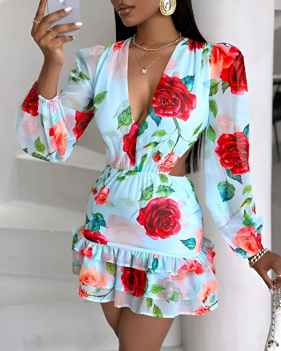 Une femme porte une robe bleu clair à motifs de roses rouges. La robe est courte et possède des manches longues. Elle est cintrée à la taille et possède un décolleté en V. La femme est debout et tient un téléphone portable à la main.