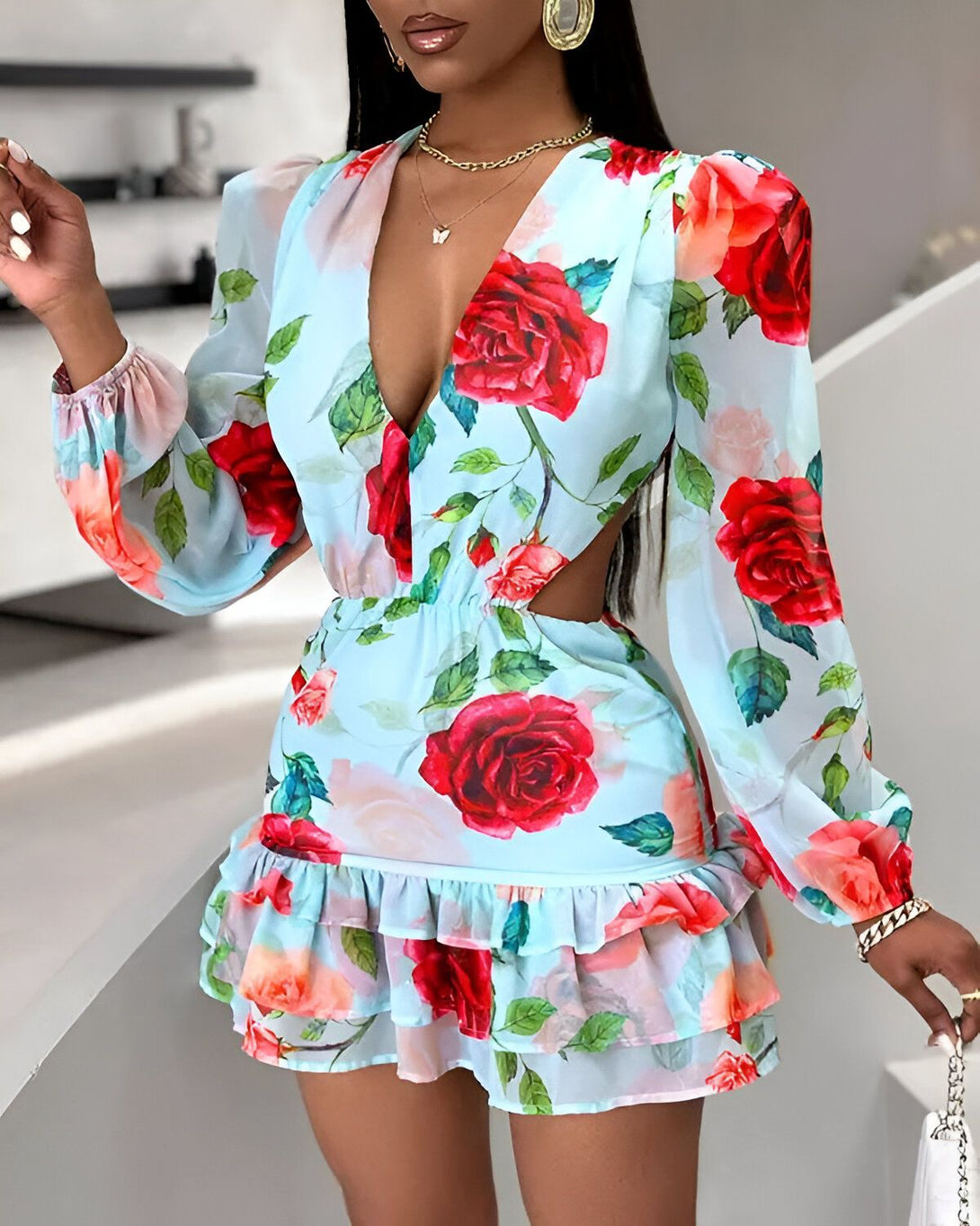 Une femme porte une robe bleu clair à motif de roses rouges. La robe est courte et a un décolleté plongeant. Les manches sont longues et bouffantes. La robe est cintrée à la taille et évasée au niveau des hanches. La femme porte des escarpins blancs et un collier doré. Elle a les cheveux longs et noirs et le maquillage léger. Elle est debout dans une pièce blanche.