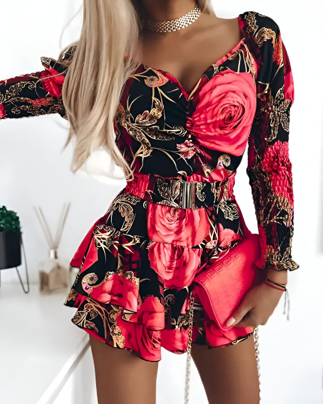 Une femme porte une combinaison short rouge et noir à motifs floraux. La combinaison est cintrée à la taille et possède un décolleté en V. Les manches sont longues et bouffantes. La femme porte également un collier doré et un sac à main rouge.