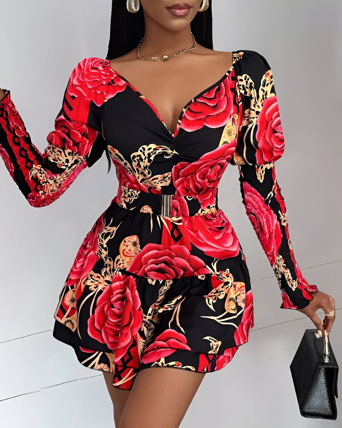 Une femme porte une robe noire à fleurs rouges. La robe est courte et cintrée à la taille. Elle a des manches longues et un col en V. La femme porte également un collier et des boucles d'oreilles. Elle a les cheveux longs et noirs et un maquillage léger. Elle porte un sac à main noir à la main.