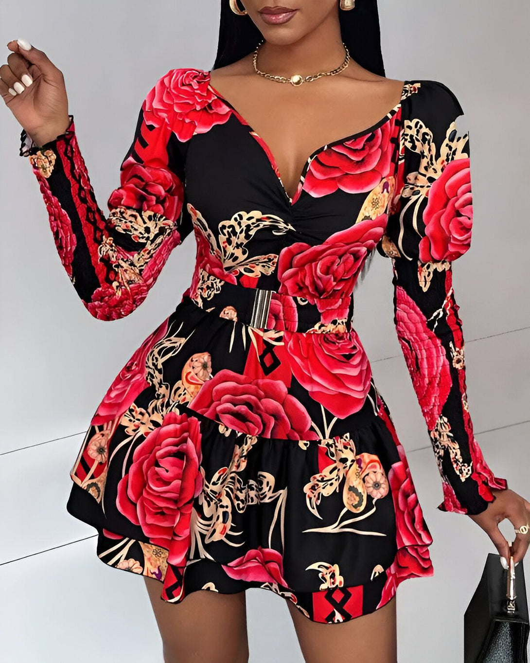 Une femme porte une robe noire et rouge à motifs floraux. La robe est courte et évasée, avec un décolleté en V et des manches longues. La femme est debout, avec une main sur la hanche et l'autre tenant un sac à main noir.