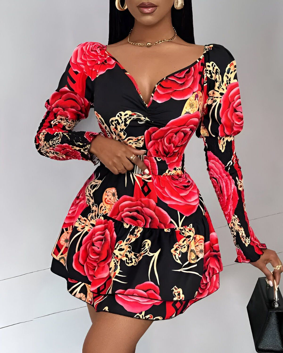Une femme porte une robe noire et rouge à motifs floraux. La robe est courte et évasée, avec un décolleté en V et des manches longues. La femme est également porte une ceinture noire et des escarpins noirs. Elle a les cheveux longs et noirs et porte un maquillage léger.