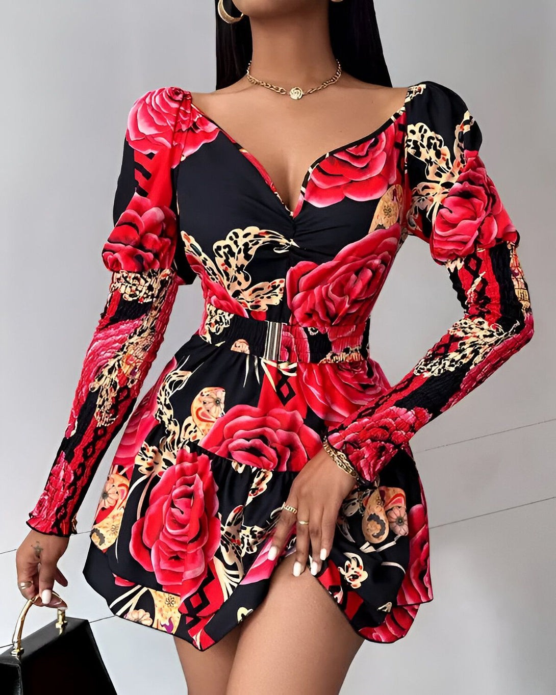 Une femme porte une robe noire et rouge à motifs de roses. La robe est courte et cintrée à la taille. Elle a des manches longues et un col en V. La femme est debout et porte des talons noirs. Elle a les cheveux longs et noirs et un maquillage léger.
