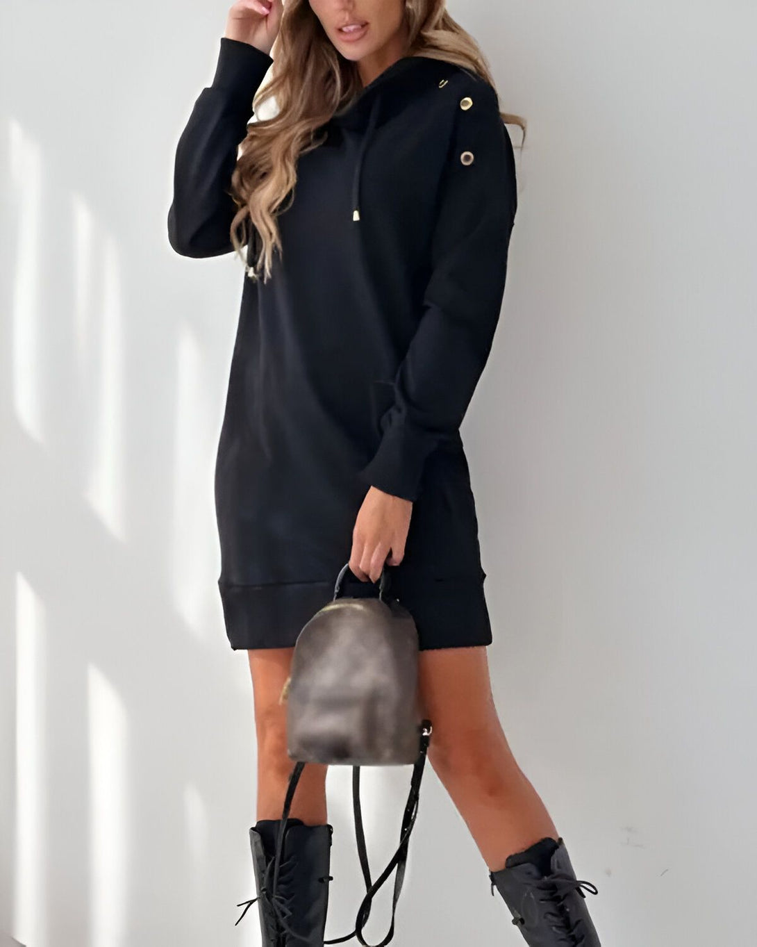 Une jeune femme porte une robe pull noire à capuche et des bottes noires. Elle porte également un sac à dos marron.
