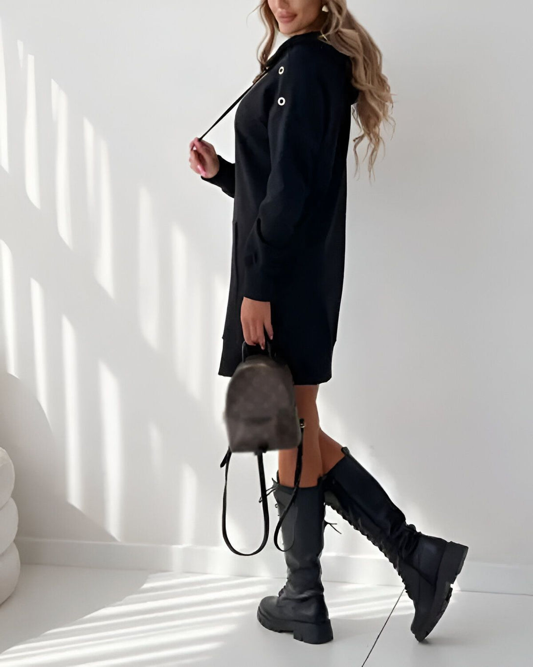 Une femme porte une robe noire à capuche et des bottes noires. Elle a un sac à dos marron.