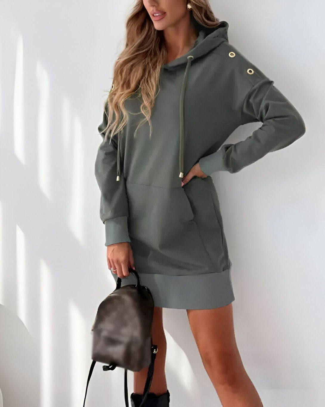 Une jeune femme porte une robe pull à capuche verte. La robe est mi-longue et possède une capuche avec des boutons dorés. La femme porte également un sac à dos marron et des bottes noires.