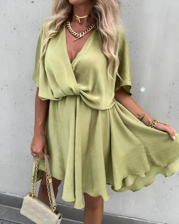 Une femme porte une robe verte courte en satin. La robe a un décolleté en V et des manches courtes. La femme porte également un collier et un sac à main.