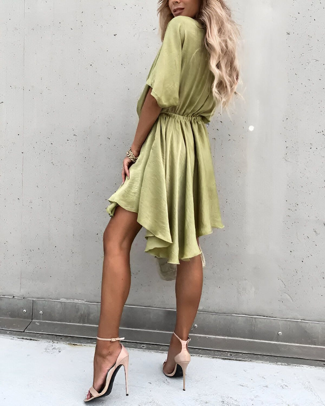 Une femme porte une robe verte courte en satin. La robe est froncée à la taille et possède un décolleté en V. Elle est associée à des sandales à talons hauts de couleur beige.