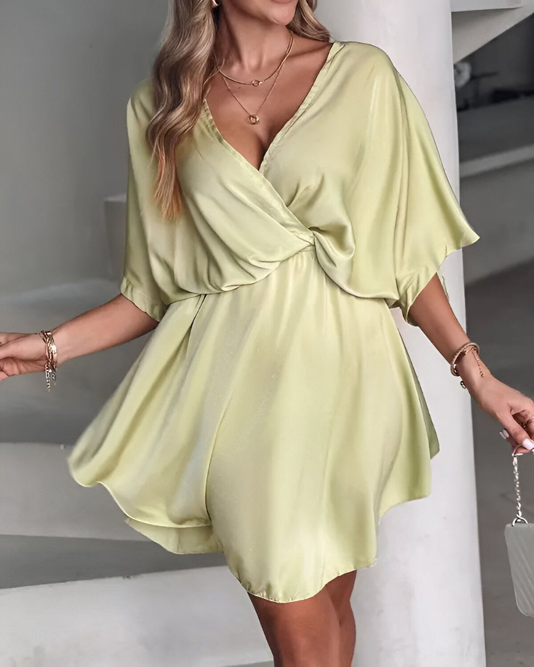 Une femme porte une robe verte courte en satin. La robe est décolletée en V et a des manches courtes. La femme est blonde et porte des bijoux dorés. Elle est debout sur un fond blanc.