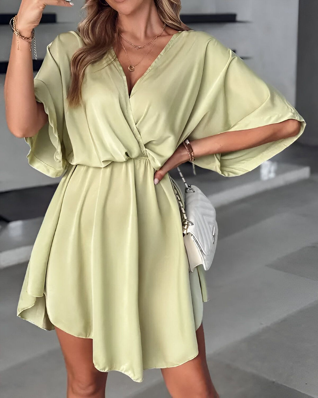 Une jeune femme porte une robe verte courte en satin. La robe est décolletée en V et a des manches courtes. La femme est blonde et porte des talons hauts blancs. Elle a un sac à main blanc à la main droite.