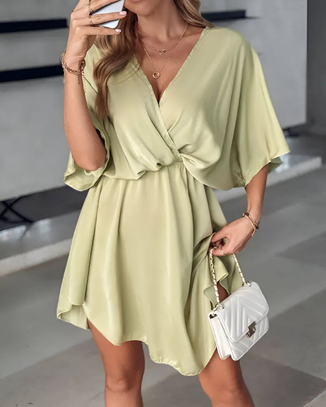 Une femme porte une robe verte satinée décolletée et des manches courtes. Elle porte également un collier et un bracelet. Elle tient un sac à main blanc à la main.