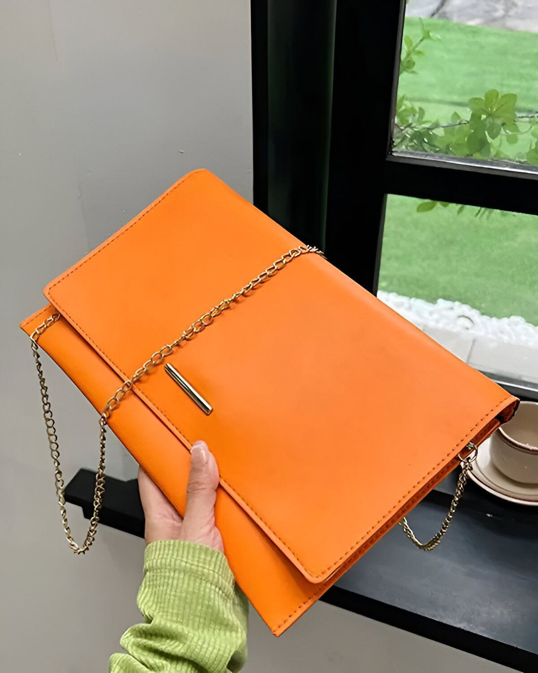 Une femme tient un sac à main orange vif à la main. Le sac à main est en cuir et possède une chaîne dorée. La femme porte un pull vert.
