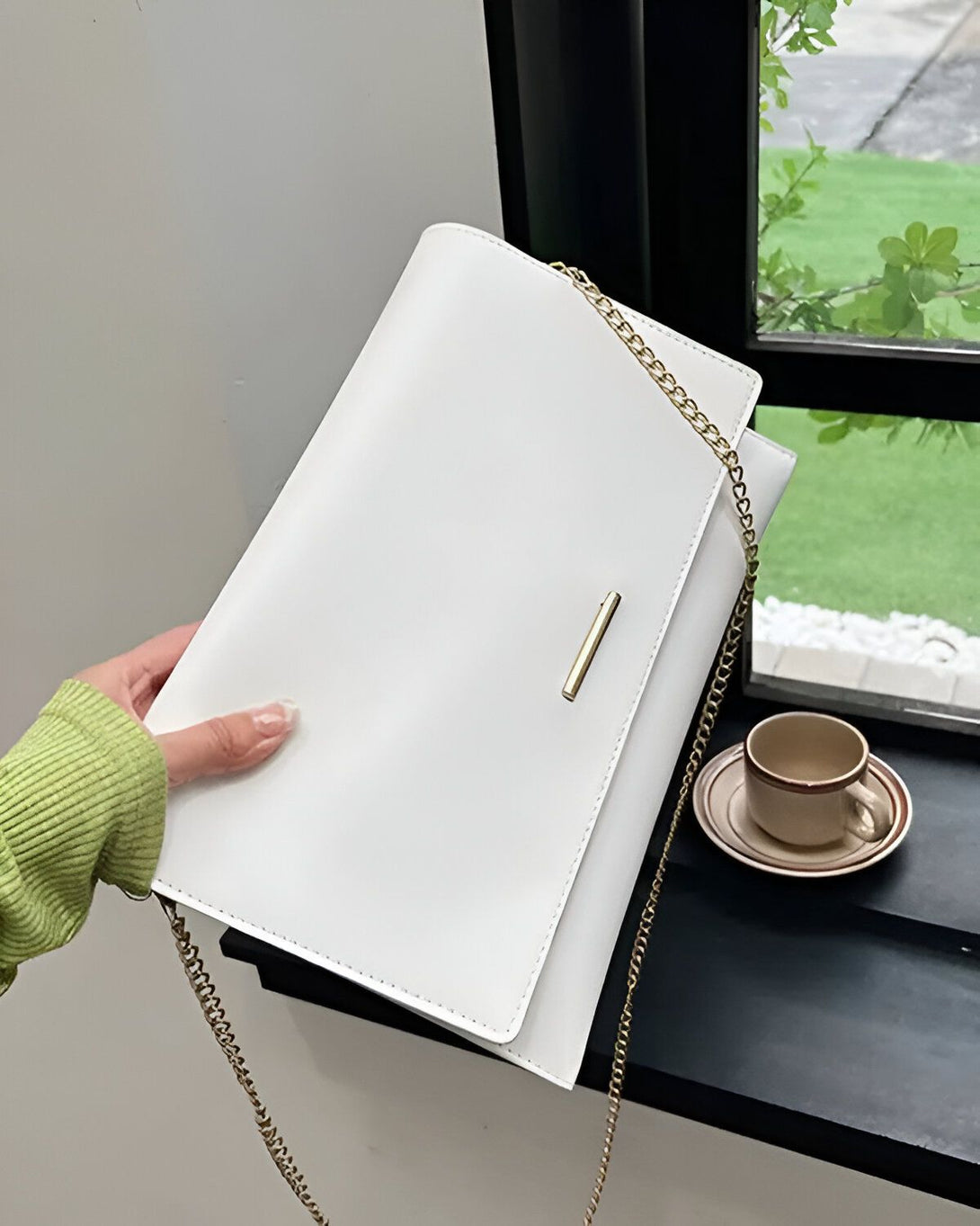 Une image d'un sac à main blanc avec une chaîne en or. Le sac est porté par une femme vêtue d'un pull vert. Le sac est placé sur une table près d'une fenêtre.