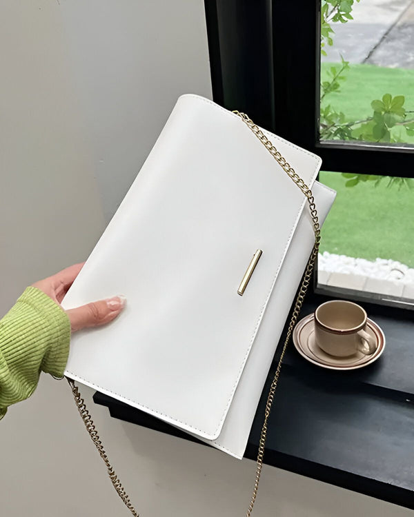 Une image d'un sac à main blanc avec une chaîne en or. Le sac est porté par une femme vêtue d'un pull vert. Le sac est placé sur une table près d'une fenêtre.