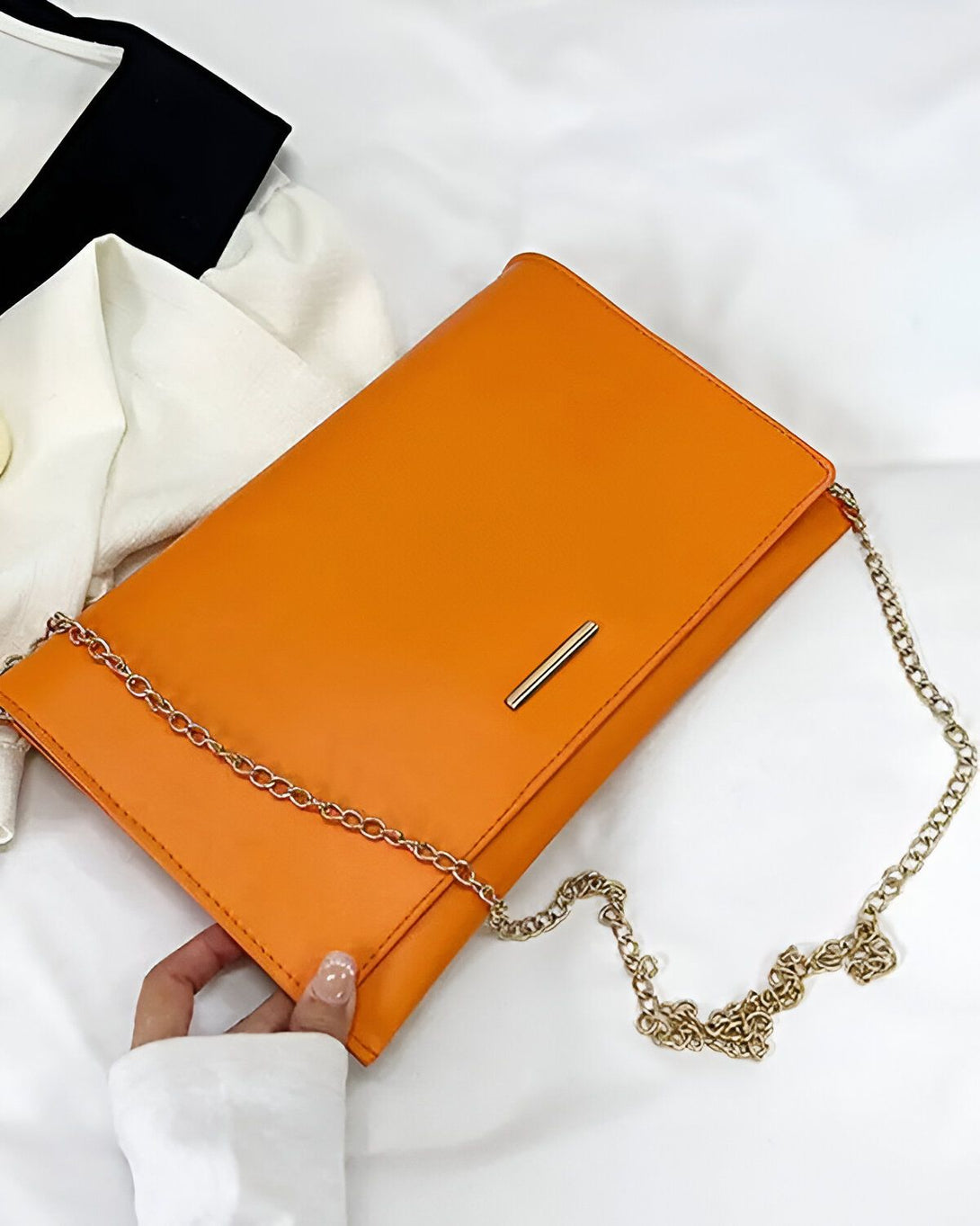 Une image d'un sac à main orange avec une chaîne dorée. Le sac à main est porté par une femme vêtue d'une chemise blanche. Le sac à main est en cuir et a une forme rectangulaire. La chaîne est en métal doré et est attachée au sac à main par deux mousquetons. Le sac à main est ouvert et on peut voir qu'il est doublé de tissu orange.