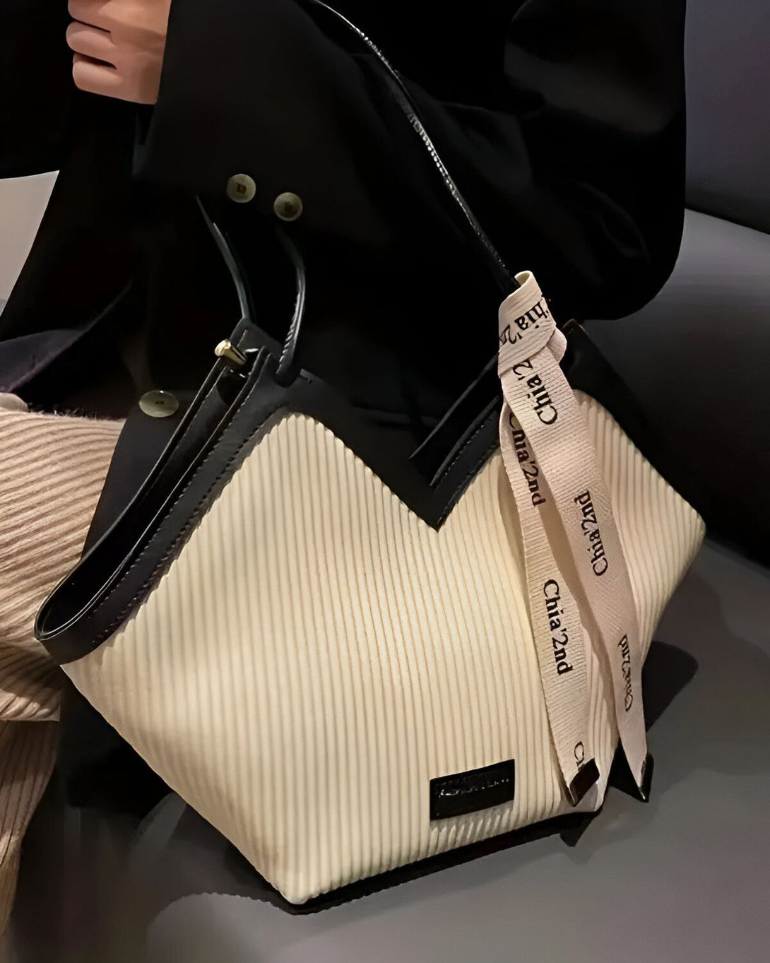 Une femme porte un manteau noir et un pantalon beige. Elle tient un sac à main blanc et beige à la main. Le sac à main est de forme trapèze et possède une anse noire. Il est décoré d'un ruban beige avec le logo "Chia 2nd" écrit en noir.