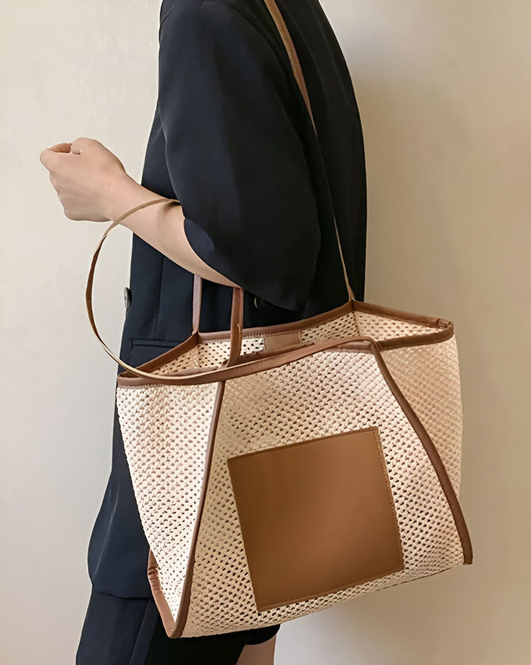 Une femme porte un cabas beige et marron en bandoulière. Le cabas est en matière tressée et possède une grande anse. La femme est vêtue d'un tailleur noir.