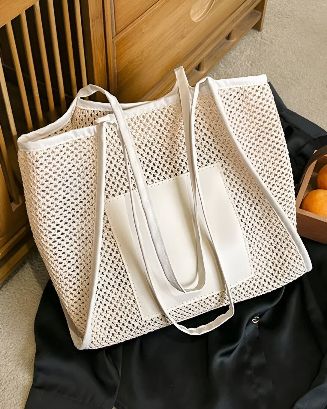 Un sac fourre-tout en filet beige avec des anses blanches et une poche avant blanche. Il est posé sur un sol en bois.