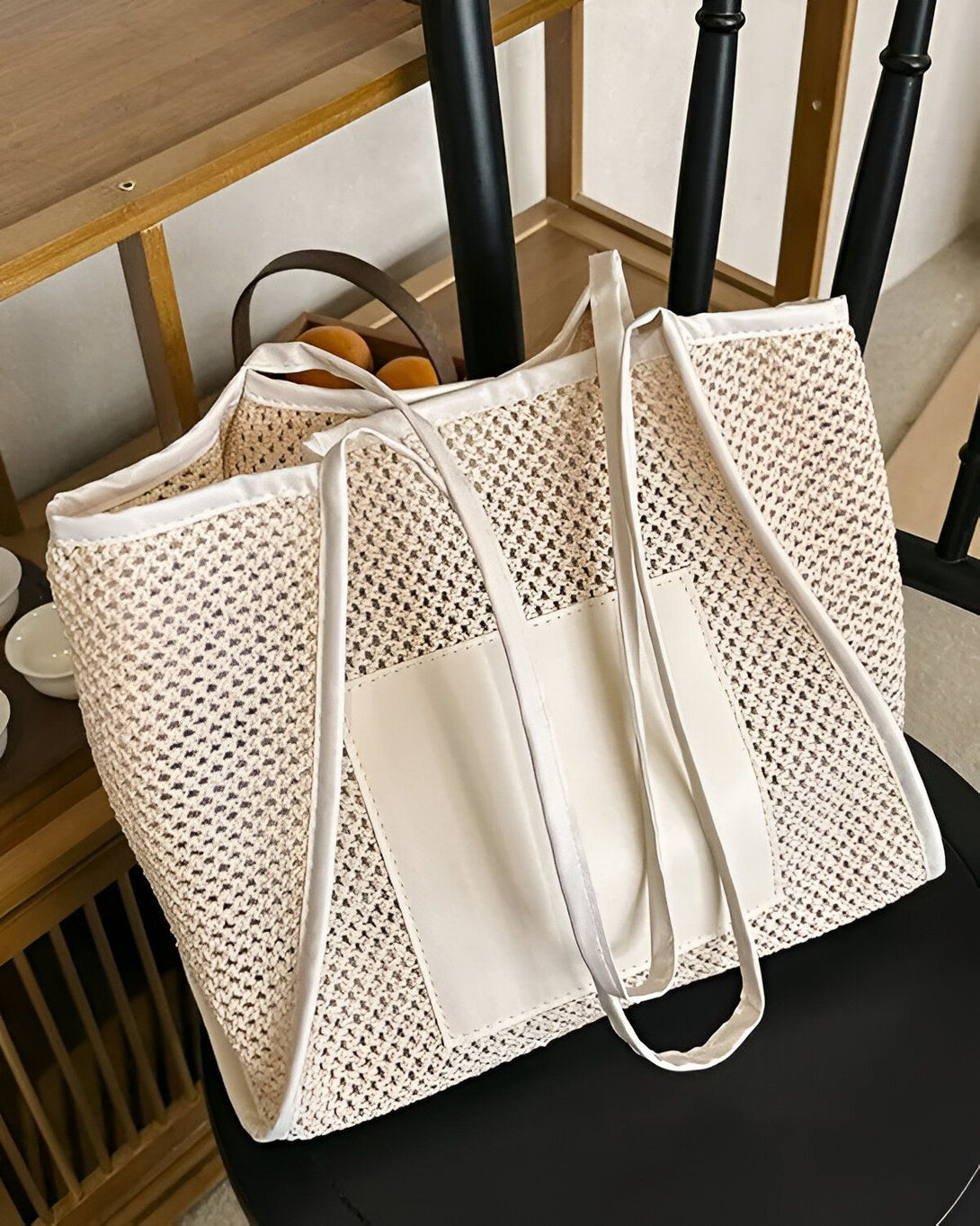 Un sac fourre-tout en filet beige avec des anses blanches et une poche avant blanche. Il est posé sur une chaise en bois.