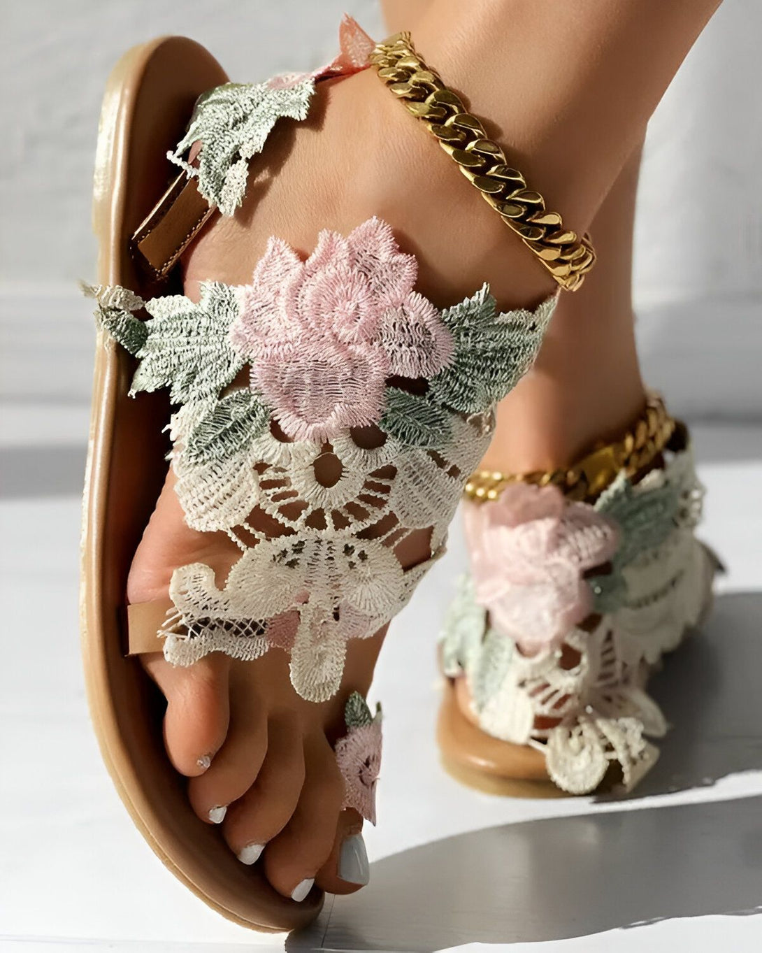 Une femme porte des sandales plates marron clair avec des fleurs en dentelle rose et blanche. Les sandales sont attachées à la cheville avec une chaîne en or.