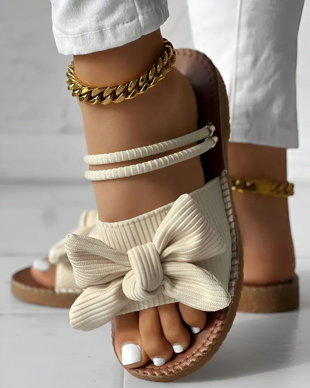 Une femme porte des sandales beiges à talons compensés. Les sandales sont ornées d'un nœud sur le dessus. La femme porte également un pantalon blanc et un bracelet de cheville doré.