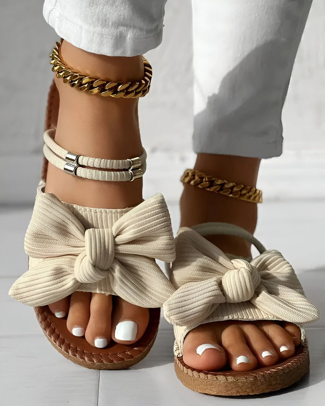 Une femme porte des sandales beiges à talons compensés. Les sandales sont ornées d'un nœud sur le dessus. La femme porte également un pantalon blanc et des bijoux dorés.
