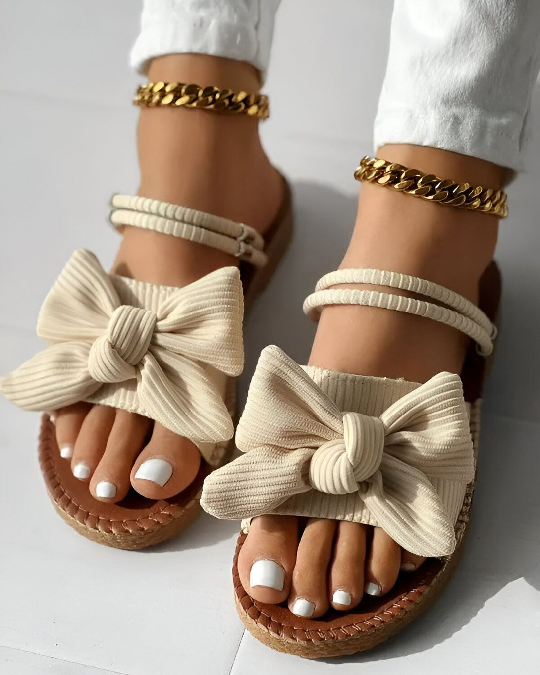 Une femme porte des sandales plates beiges avec un nœud sur le devant. Les sandales sont en tissu et ont une semelle en corde. La femme a les ongles des pieds peints en blanc et porte des bijoux de cheville en or.