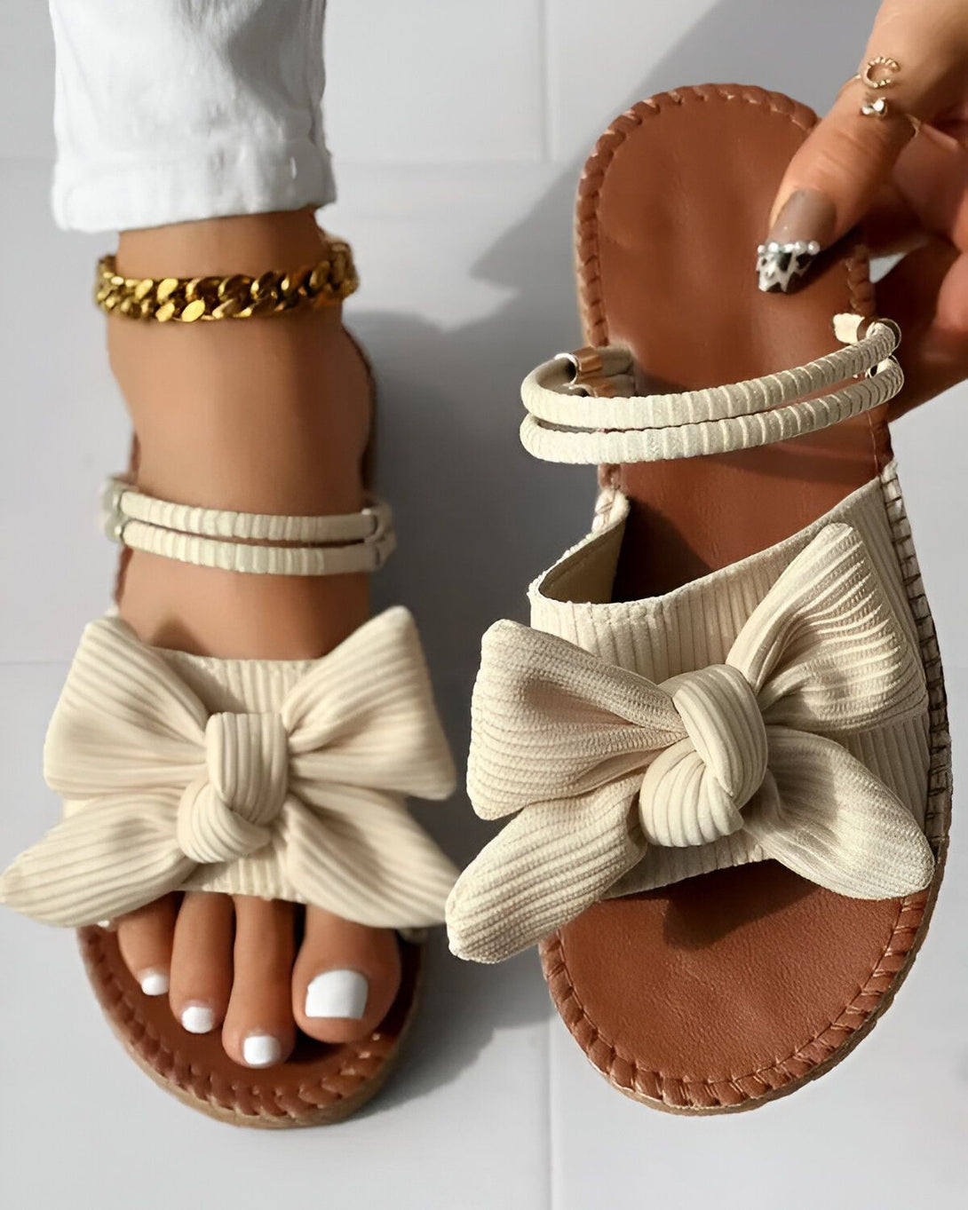 Une femme porte des sandales plates beiges avec un nœud sur le devant. Les sandales sont en tissu et ont une semelle en caoutchouc. La femme a les ongles des pieds peints en blanc et porte un bracelet de cheville doré.