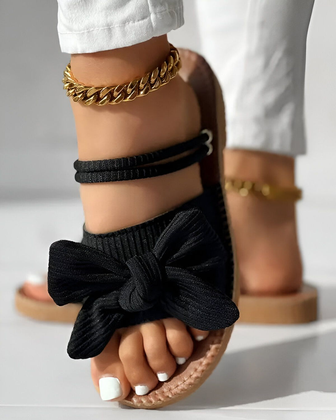 Une femme porte des sandales noires à talons compensés. Les sandales sont dotées d'un nœud en tissu noir sur le dessus et de brides noires qui s'enroulent autour de la cheville. La femme porte également un pantalon blanc et un bracelet de cheville doré.
