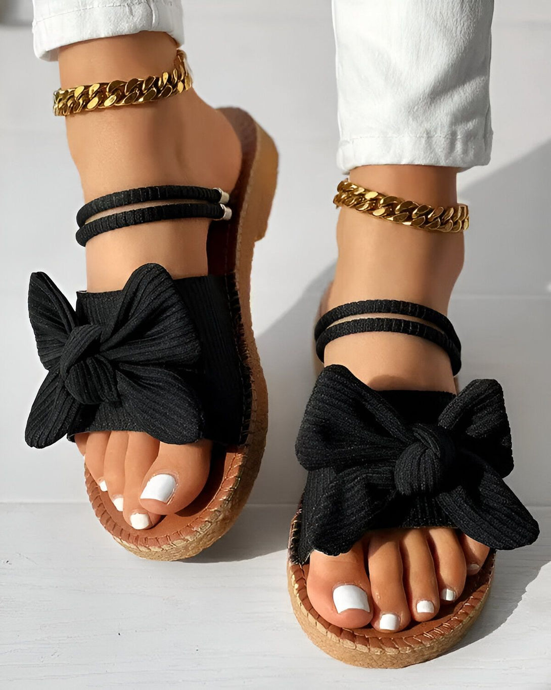Une femme porte des sandales noires à talons compensés. Les sandales sont ornées d'un nœud en tissu noir. La femme porte également un pantalon blanc et des bijoux dorés.