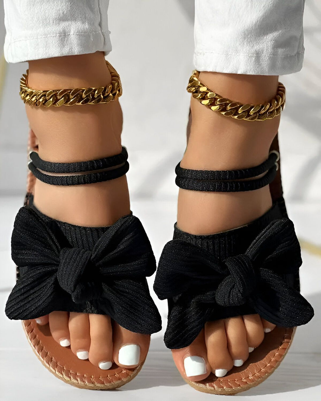 Une femme porte des sandales noires à talons compensés. Les sandales sont dotées d'un nœud en tissu sur le devant et de brides fines qui s'enroulent autour de la cheville. La femme porte également un pantalon blanc et des bracelets de cheville en or.