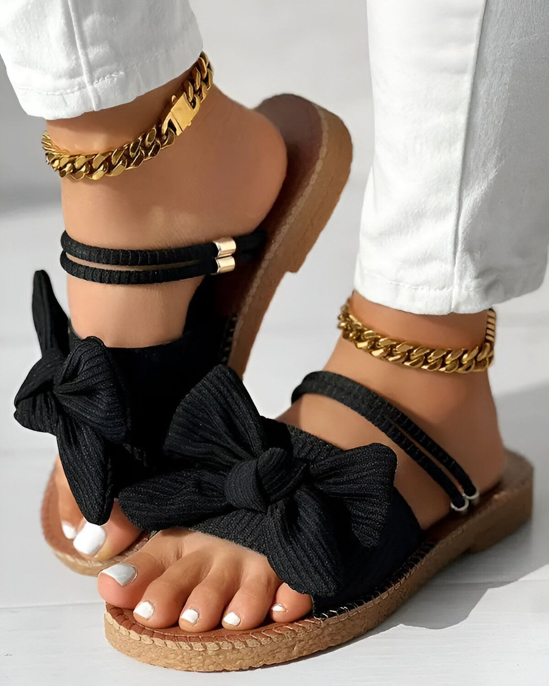 Une femme porte des sandales noires à talons compensés. Les sandales sont ornées d'un nœud en tissu noir. La femme porte également un pantalon blanc et des bijoux dorés.