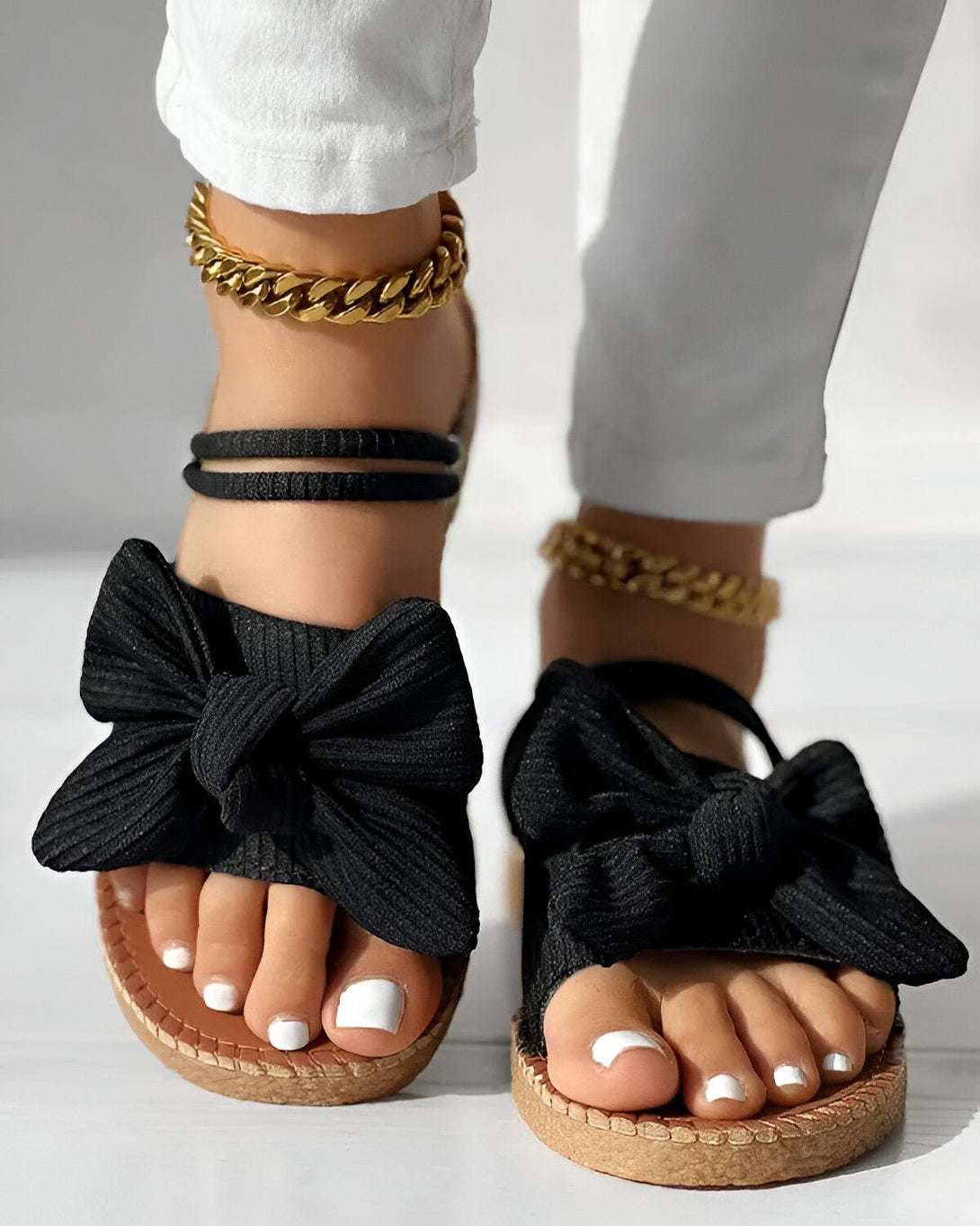 Une femme porte des sandales noires à talons compensés. Les sandales sont ornées d'un nœud en tissu noir. La femme porte également un pantalon blanc et des bijoux dorés.