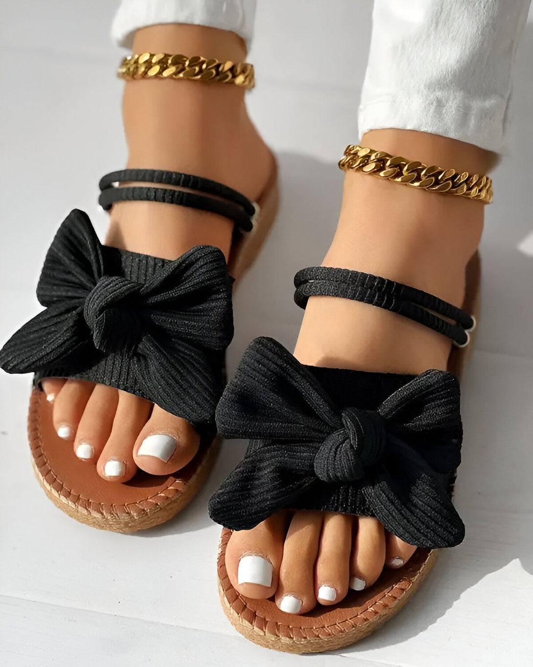 Une femme porte des sandales noires à talons compensés. Les sandales sont ornées d'un nœud en tissu noir. La femme a les ongles des pieds peints en blanc. Elle porte également un bracelet de cheville en or.