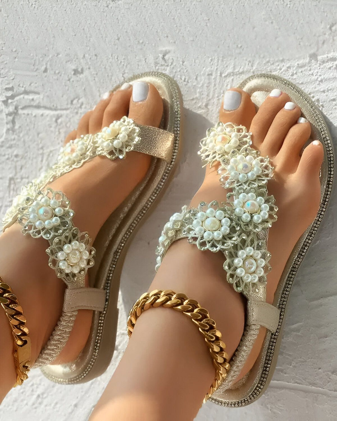 Une paire de sandales plates dorées avec des fleurs en perles blanches et des feuilles vertes. Les sandales sont portées par une femme avec une peau bronzée et des ongles blancs. La femme est debout sur un sol en béton beige.