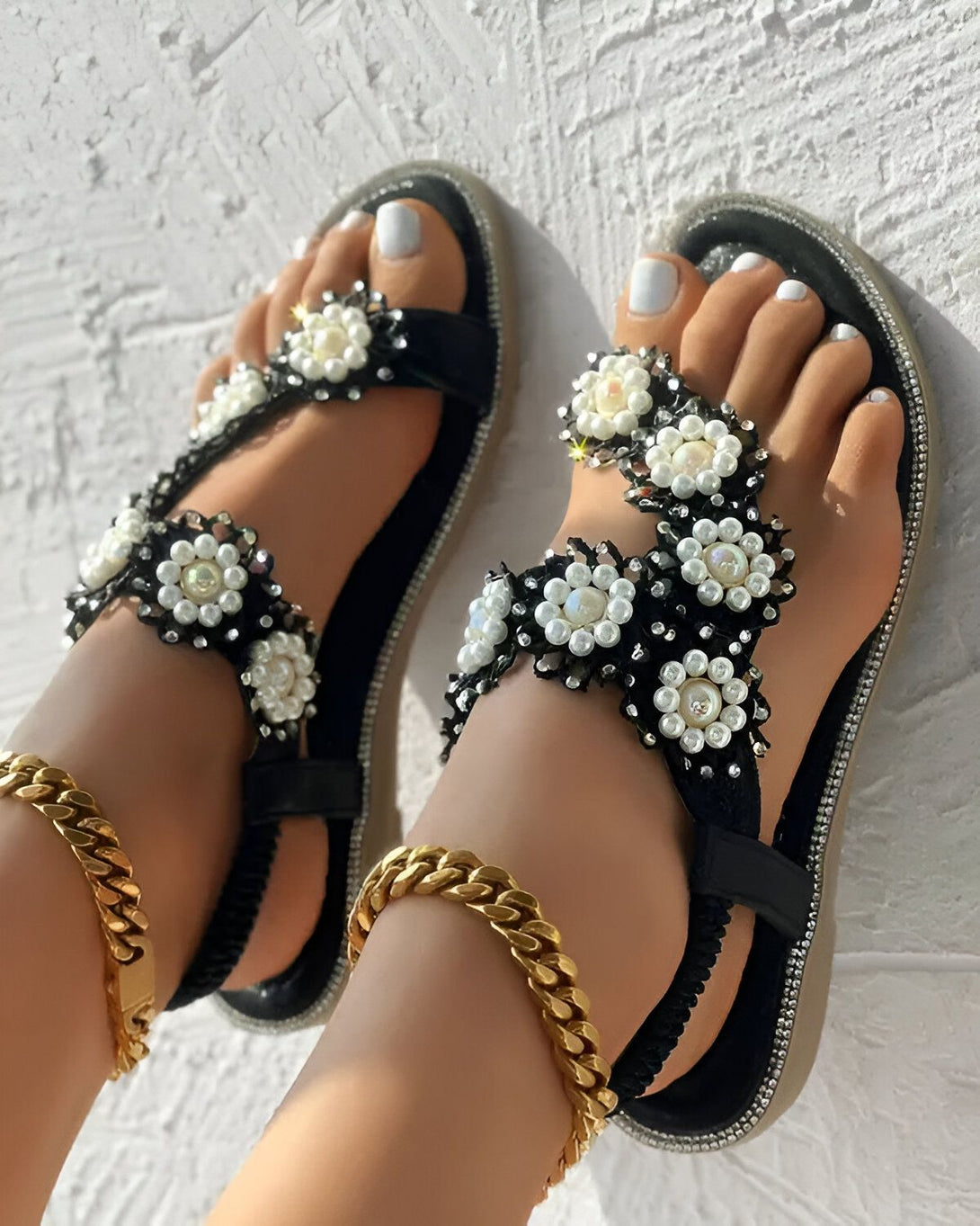 Une paire de sandales plates noires avec des fleurs en perles blanches et noires. Les sandales sont portées par une femme qui a les ongles des pieds peints en blanc. La femme est debout sur un sol en béton.