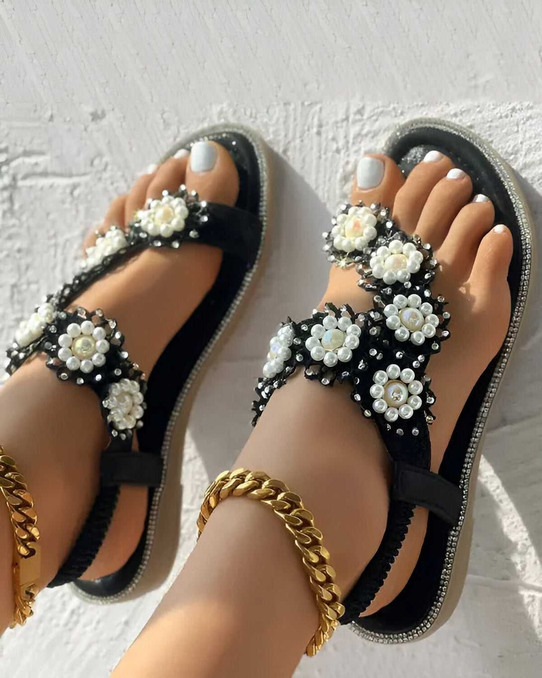 Une paire de sandales plates noires avec des fleurs en perles blanches et noires. Les sandales sont portées par un mannequin avec une peau bronzée et des ongles blancs. Le mannequin porte également un bracelet de cheville en or.
