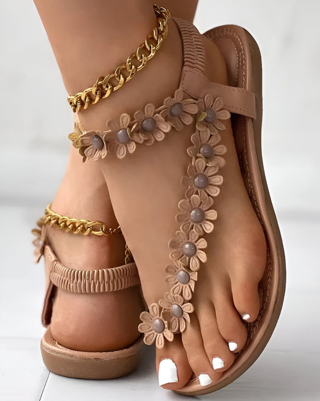 Une femme porte des sandales plates marron clair ornées de fleurs en 3D. Les sandales sont également dotées d'une chaîne dorée qui s'enroule autour de la cheville.