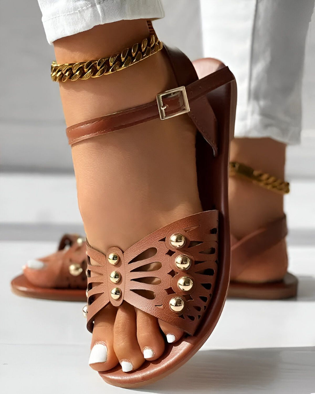 Une femme porte des sandales marron avec des clous dorés. Les sandales sont attachées avec une boucle. La femme porte également un pantalon blanc et un bracelet de cheville doré.